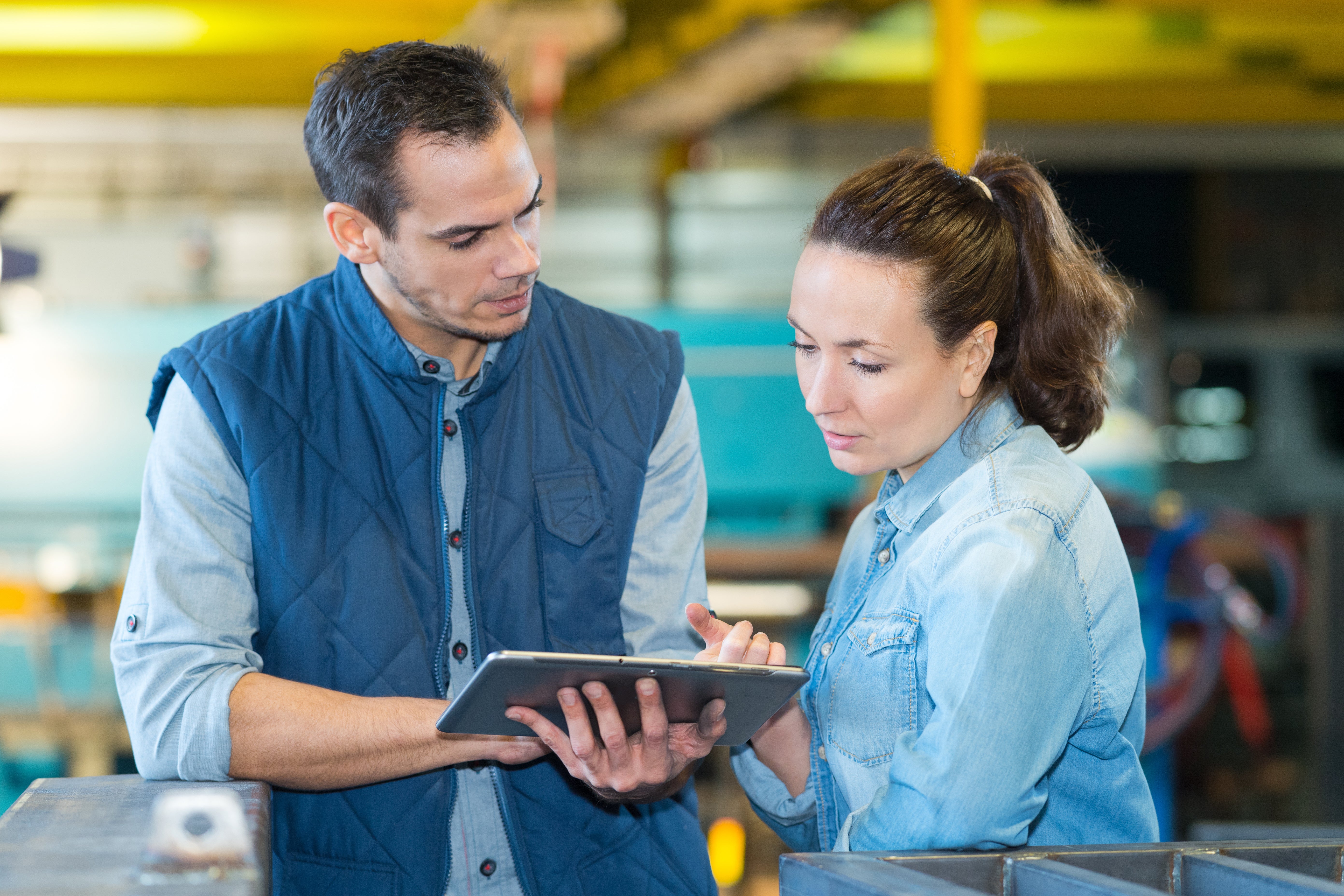 A man and women in a warehouse. They are looking at a tablet.