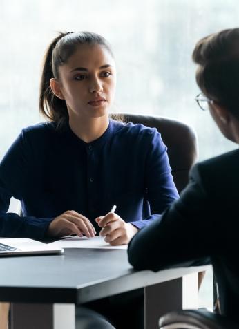 Two people facing each other, seated on opposite sides of a desk.